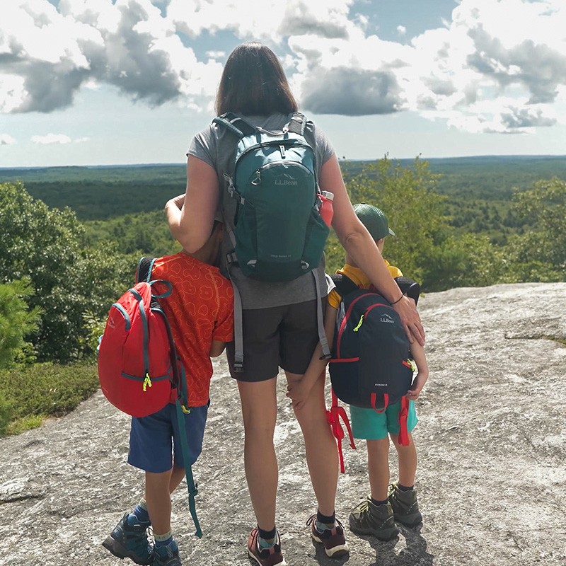 An mom and two children with stand on a rocky surface, overlooking a green landscape under a partly cloudy sky.