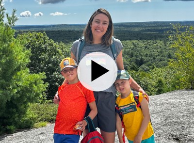An mom and two children with stand on a rocky surface, overlooking a green landscape with a play video icon in the center.