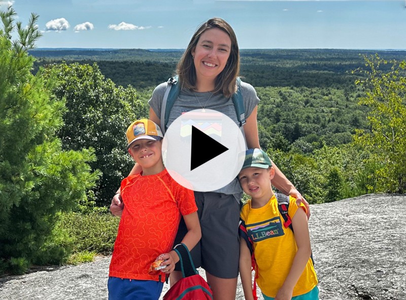 An mom and two children with stand on a rocky surface, overlooking a green landscape with a play video icon in the center.