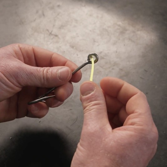 Hands threading yellow fishing line on through the eye of a fishing hook.