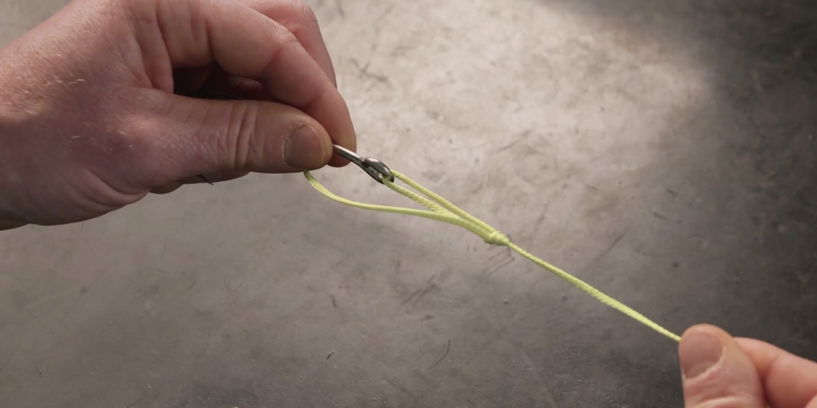 A hand holds a fishing hook that is attached to yellow fishing line via a knot. 