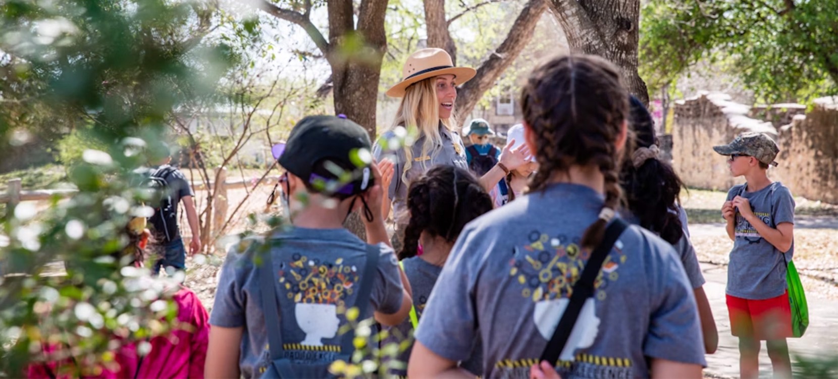 Children wearing matching shirts with a plant design on the back, gathered around an adult in a hat, outdoors near trees. 