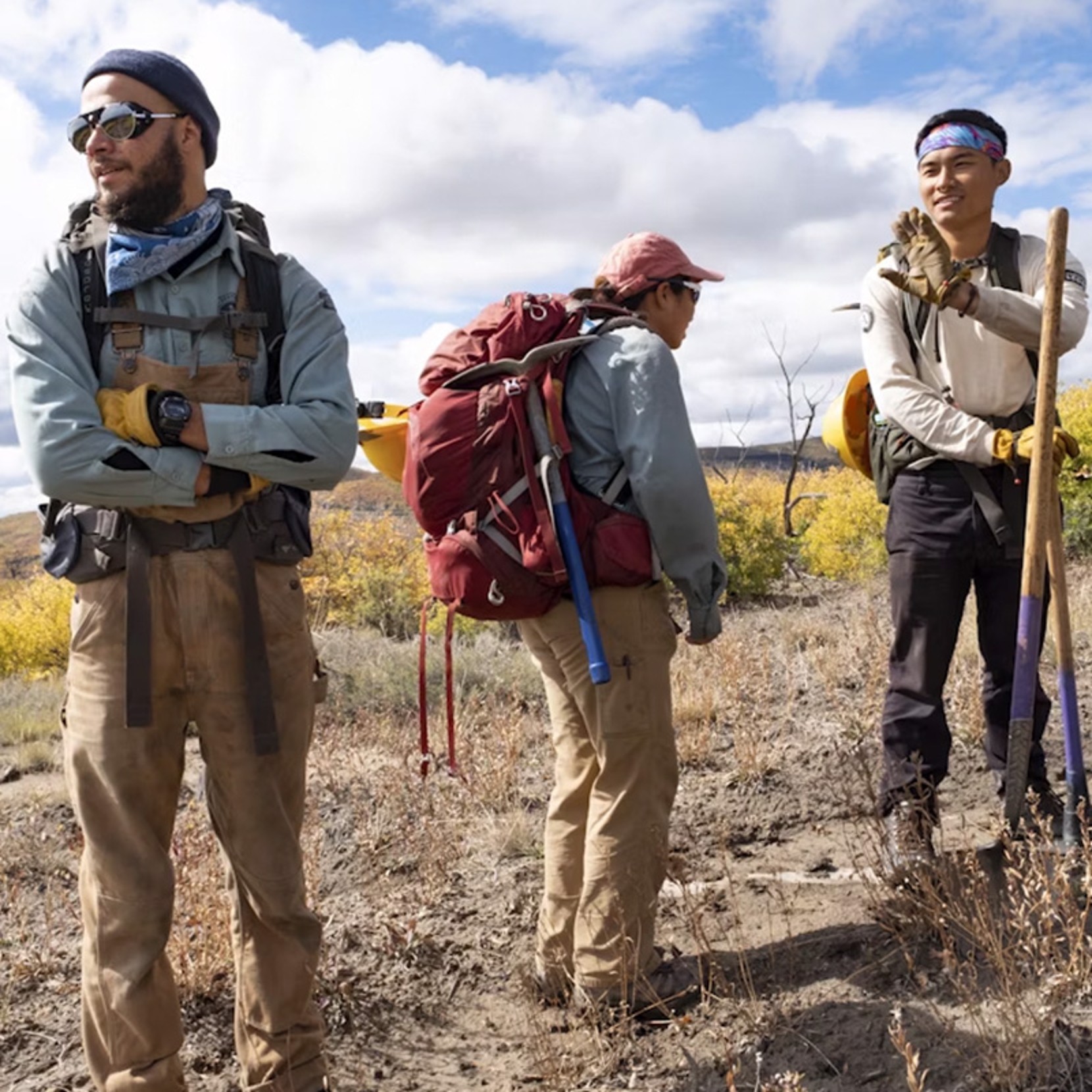 Three people in outdoor gear stand on a dirt path in a mountainous area with autumn foliage. One person holds a shovel.