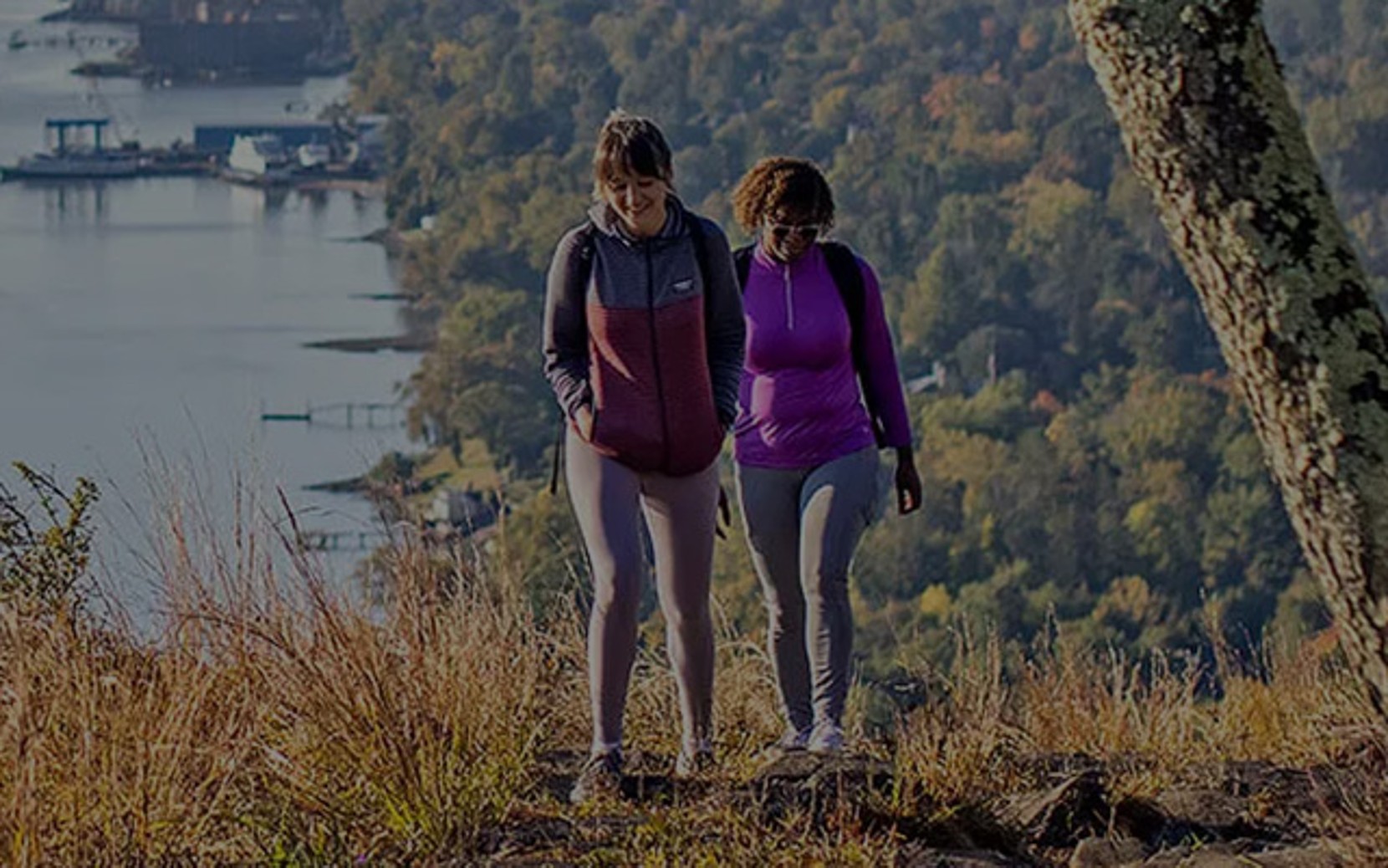 Two people hiking on a trail with a scenic view of a lake and forested hills in the background.