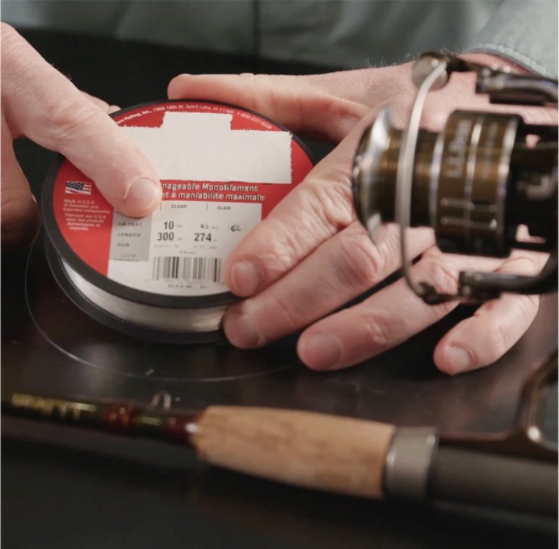 A person holds a spool of fishing line labeled "10 pounds, 300 yards, 4.4 kilograms, 274 meters" next to a fishing rod.