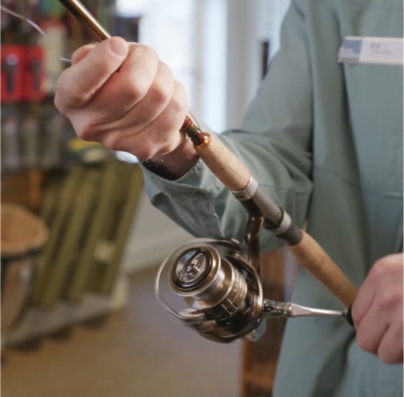 A person holding a fishing rod with a spinning reel, demonstrating how to use it in an indoor setting.