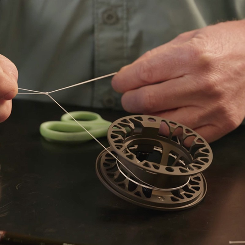 A person is threading a fishing reel with line, holding the line taut. Green scissors are on the table in the background.