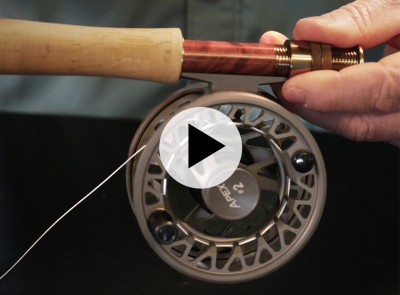 Close-up of a person holding a fishing rod with a reel labeled "APEX 2" and an overlaying play video icon.