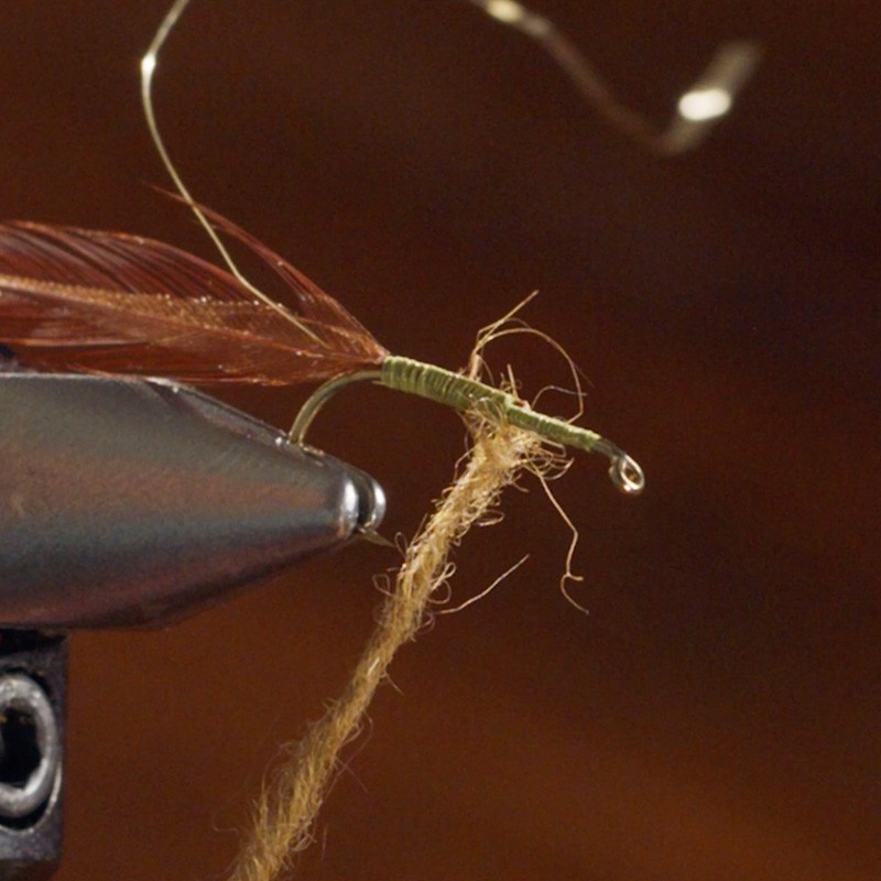A fly fishing lure being tied, with a metal vise holding the hook and various materials like feathers and thread attached.