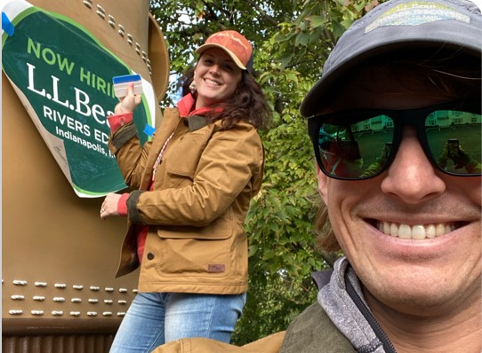 Person in a brown jacket and cap standing next to an L.L.Bean "Now Hiring" sign.