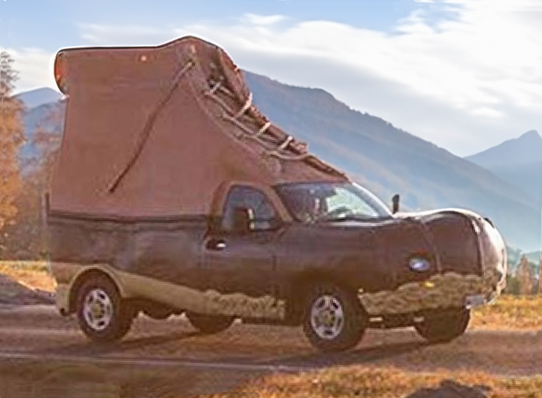 A truck modified to look like a giant L L Bean Boot with laces, driving on a road with mountains in the background.