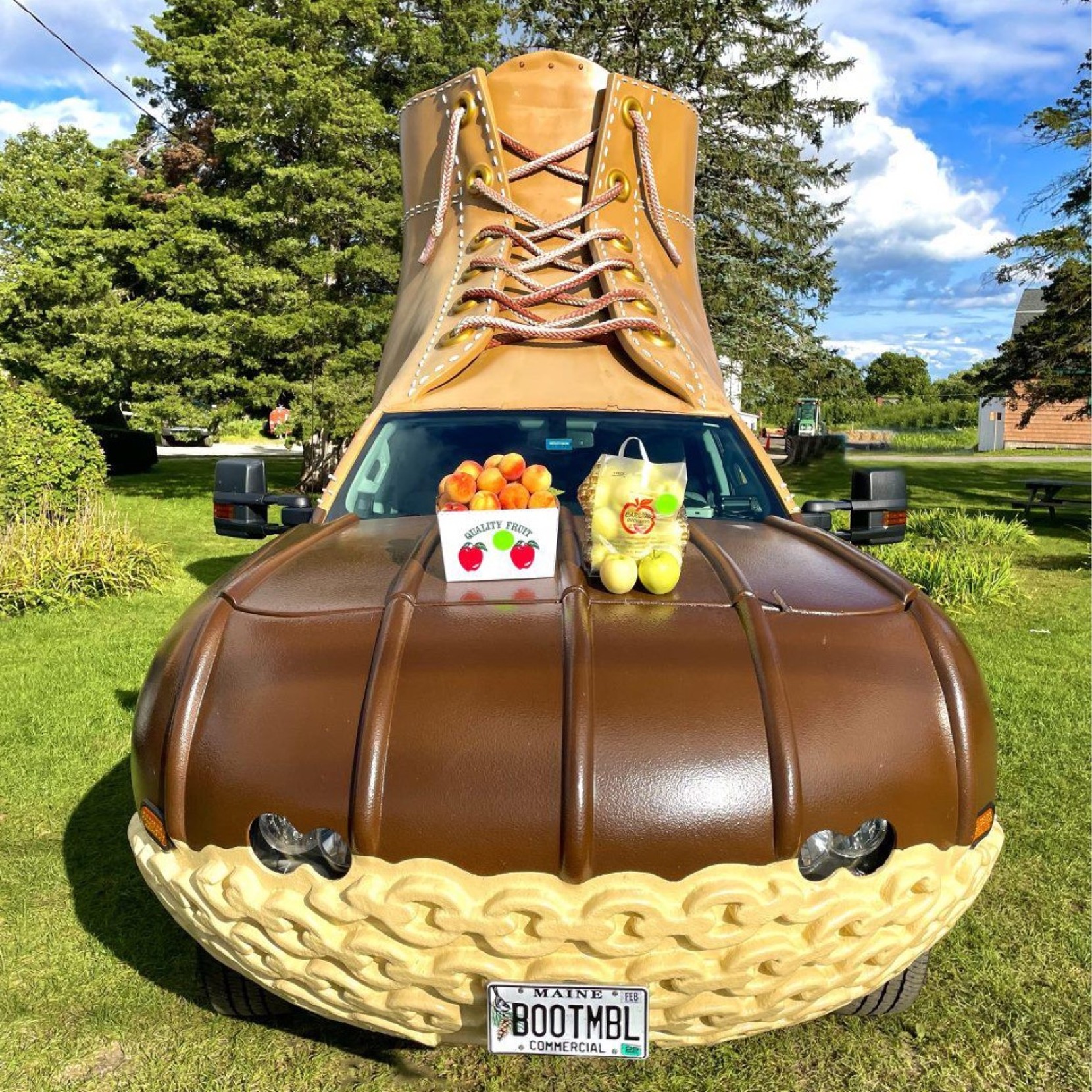A truck designed to look like a giant L L Bean Boot, holding a box of peaches and a bag of apples on the hood.