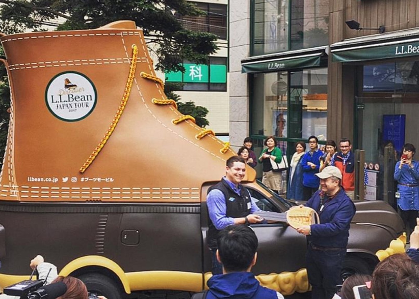 A boot-shaped vehicle is parked outside an L L Bean store, surrounded by people taking photos.