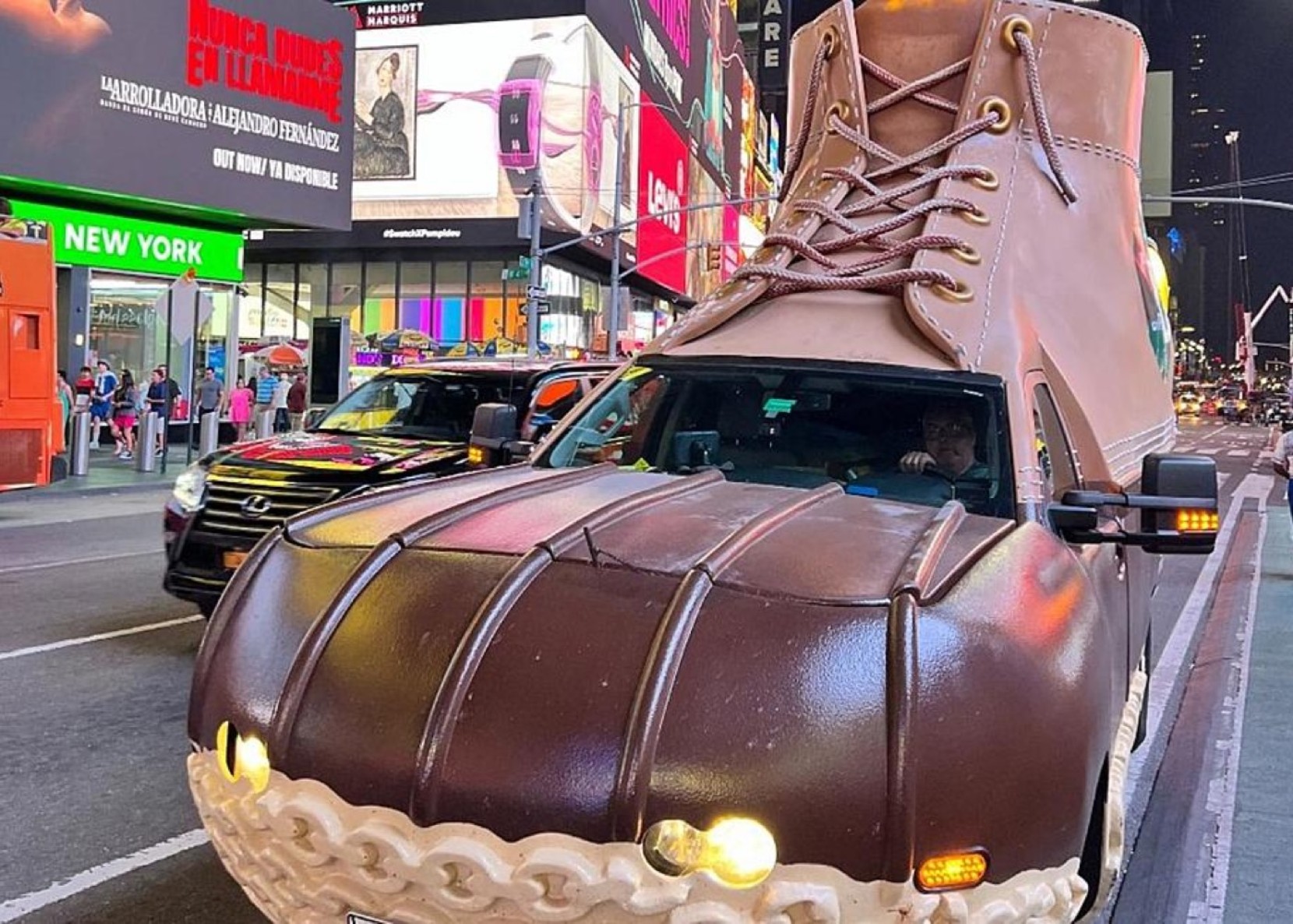 A car designed to look like a giant brown boot drives through a busy, brightly lit city street at night.
