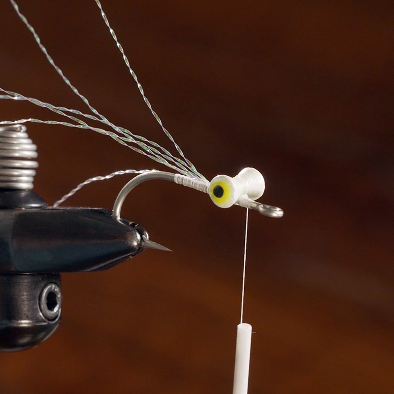 Close-up of a fishing fly being tied, showing a hook with thread and synthetic fibers attached extending from the eye.