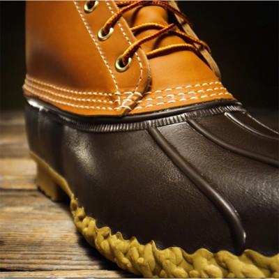 A brown and tan Bean Boot with yellow laces, detailed stitching, and a textured rubber sole on a wooden surface.