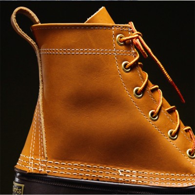 Close-up of a brown leather boot with detailed stitching, metal eyelets, and orange laces against a dark background