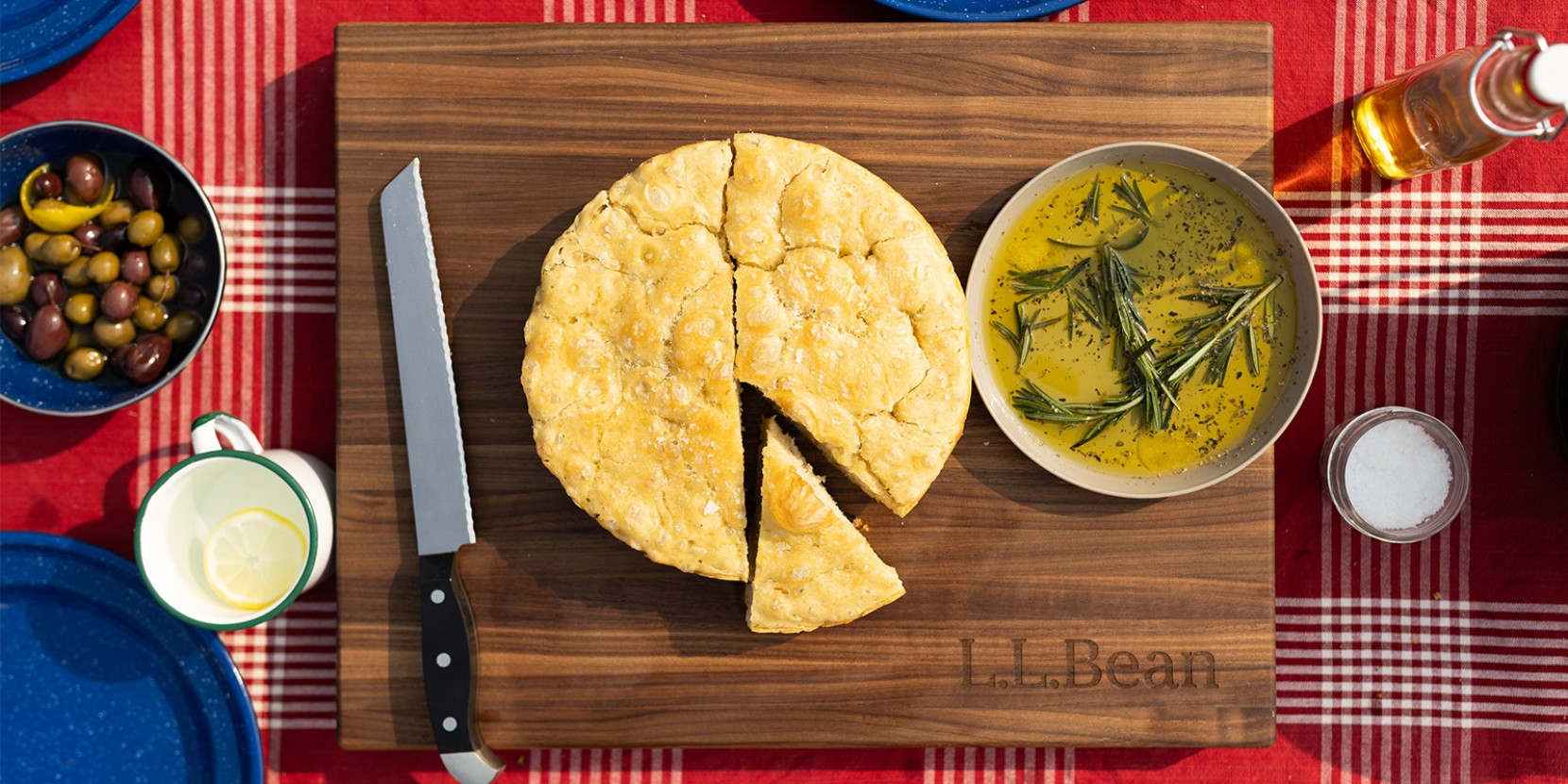 Focaccia bread with a slice cut out, knife, bowl of herbed olive oil on a cutting board board over red checkered tablecloth.