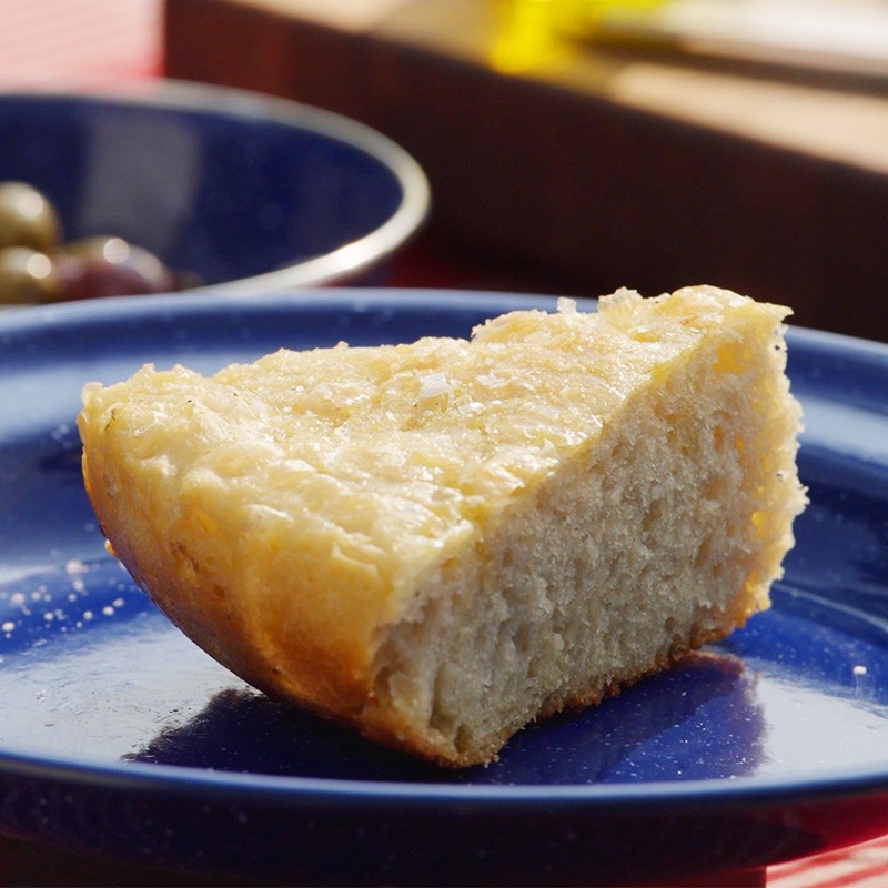 Slice of golden-crusted focaccia bread on blue plate with bowl of olives in background.