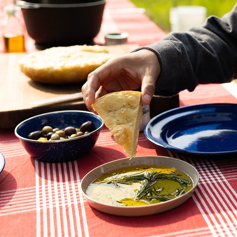 Hand dips triangular focaccia into herbed olive oil, with olives and more bread on red checkered tablecloth.