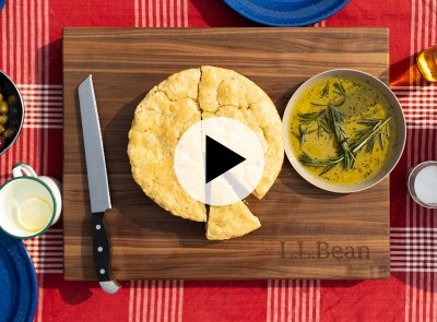 Focaccia bread on board with knife, olive oil, herbs, on red checkered tablecloth, overlaid by play video icon.