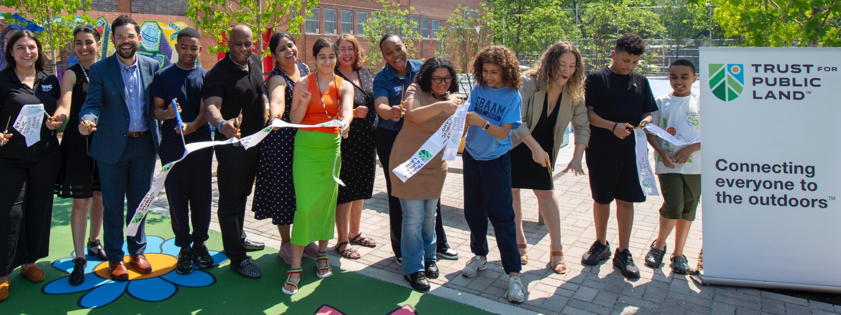 People cutting a ribbon at an outdoor event with a "Trust for Public Land" sign that reads onnecting everyone to the outdoors