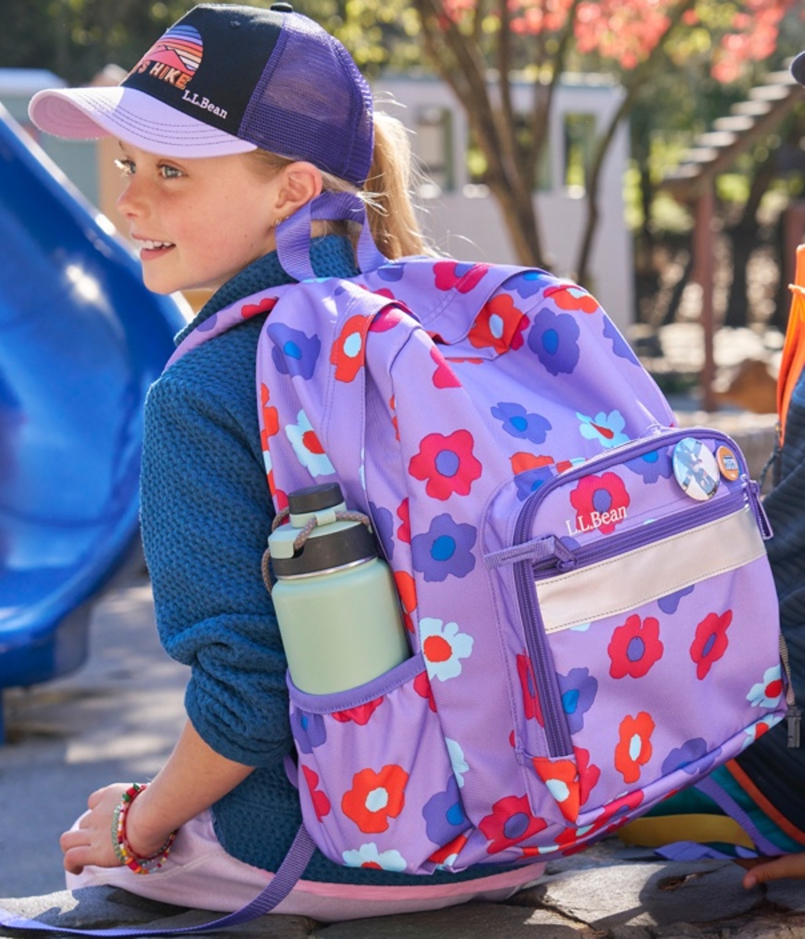 A young girl shown from behind, sitting outside and looking left wearing an L. L. Bean school backpack.