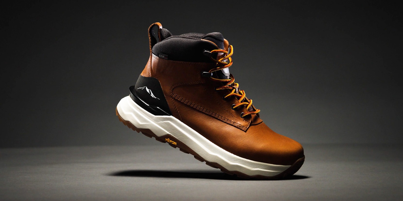 Brown leather hiking boot with orange laces, black/white accents, and thick white Vibram sole on a dark background.
