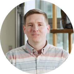 Max D in striped button-up shirt indoors, with blurred face, window, and wooden shelves in the background.
