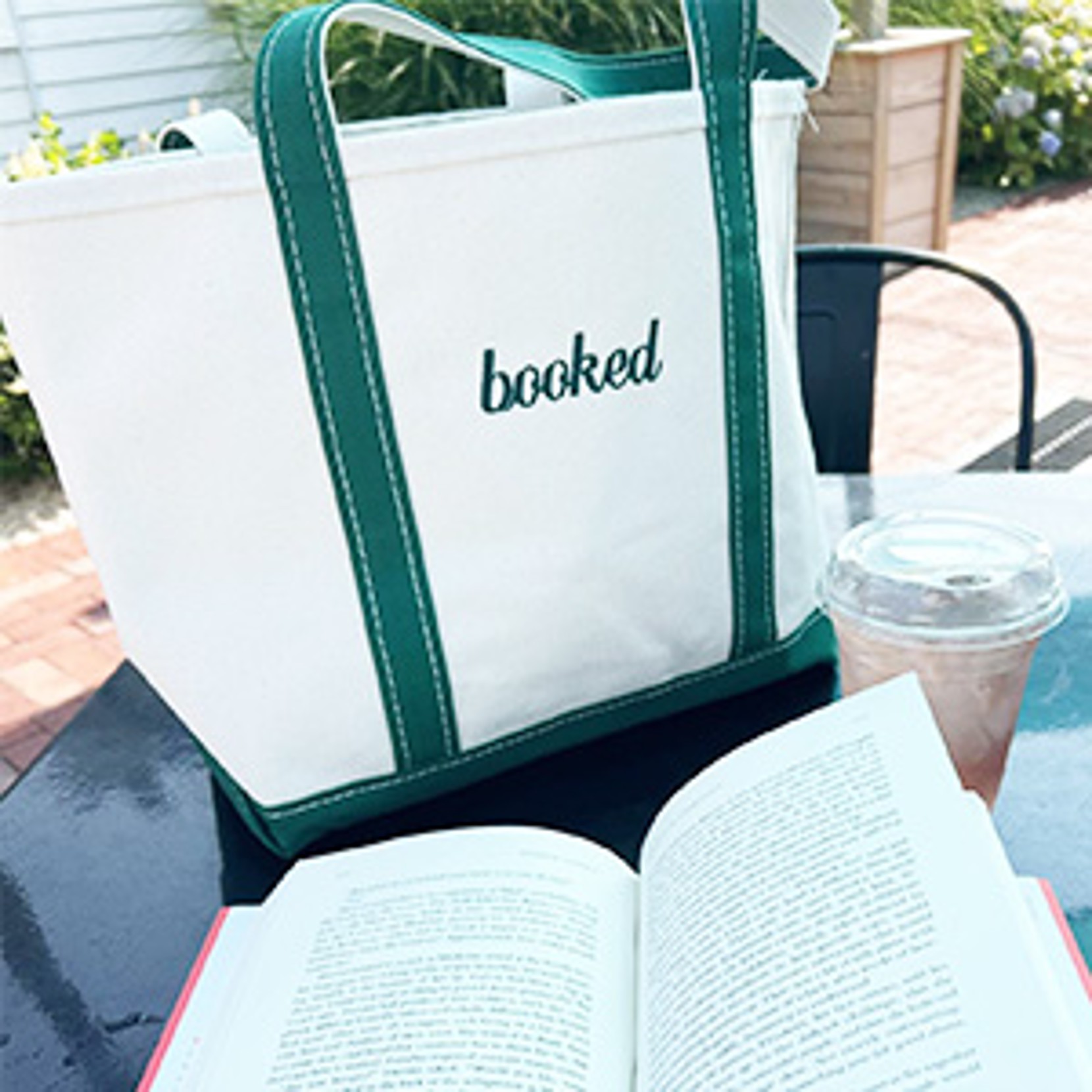 White tote with green straps labeled "booked" beside a book and iced drink on a table.