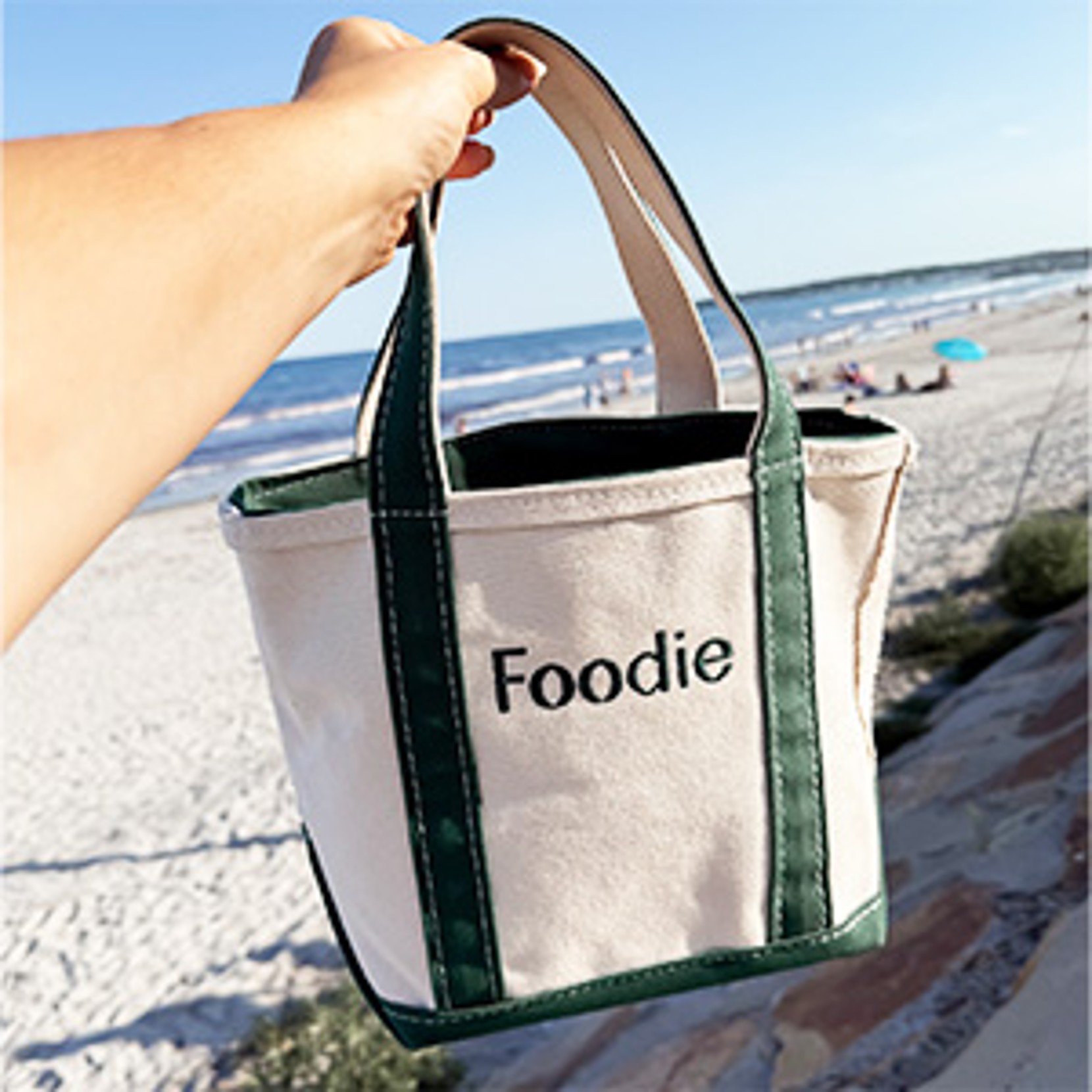 Hand holding "Foodie" tote with green straps at the beach.