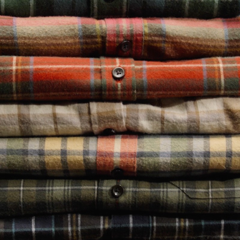 Stack of folded, flannel plaid shirts in red, green, and beige tones, neatly arranged with buttons visible.