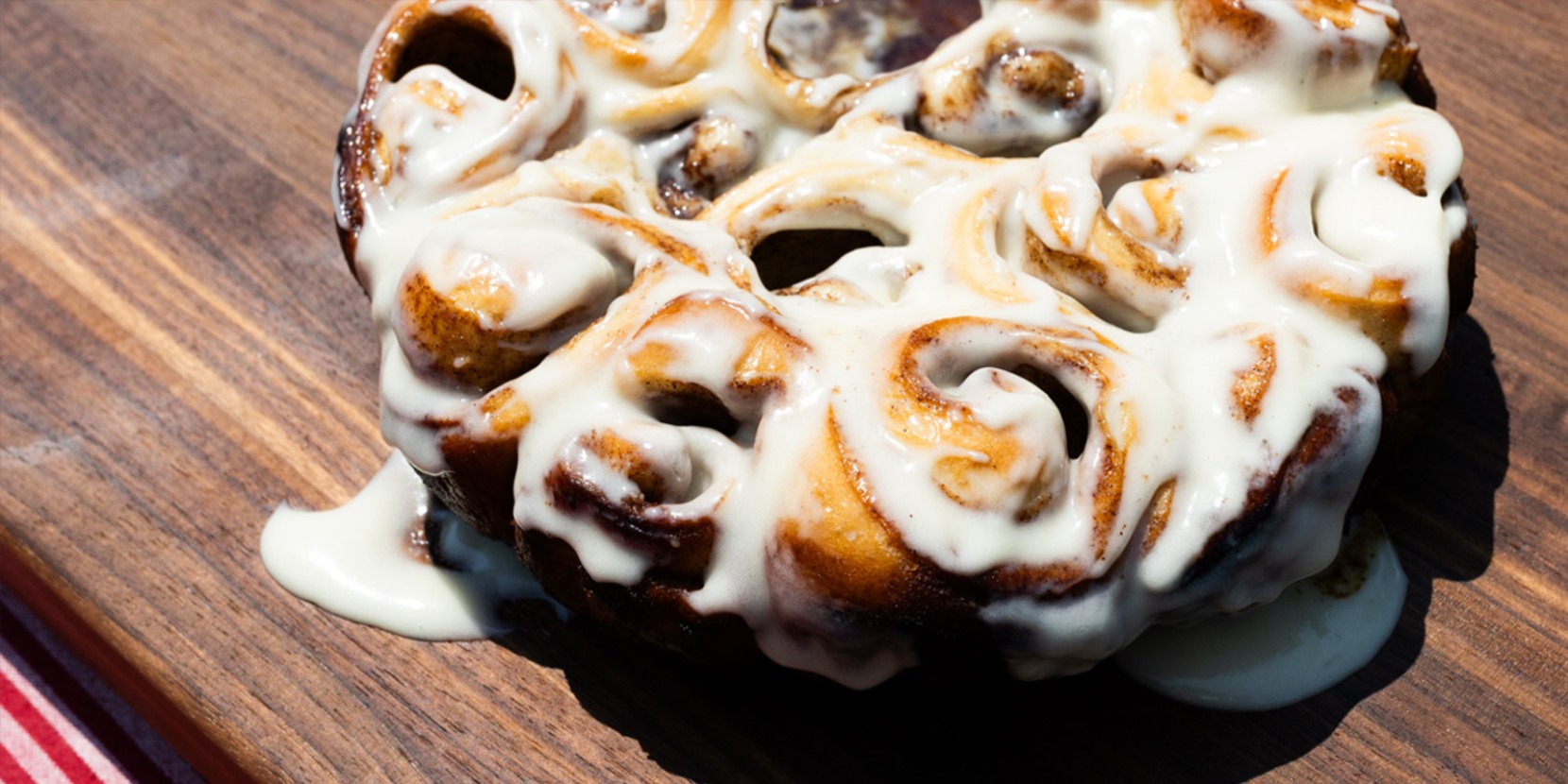 Freshly baked cinnamon rolls with white icing melting slightly on a wooden board in sunlight.