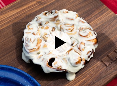 Cinnamon roll cake with white icing on a wooden board, play button overlay in center, blue plate partially visible.