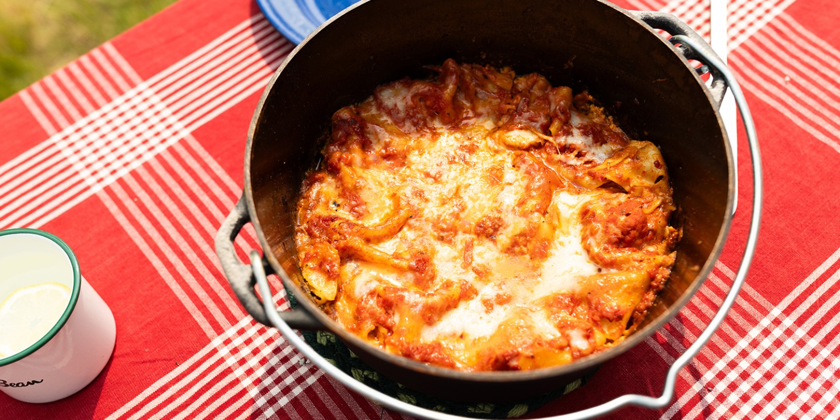 A dutch oven filled with baked lasagna with melted cheese and tomato sauce on a red checkered tablecloth.