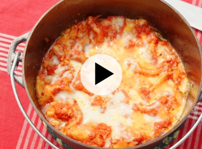 A dutch oven filled with baked lasagna with melted cheese and tomato sauce on a red checkered tablecloth with video play button.
