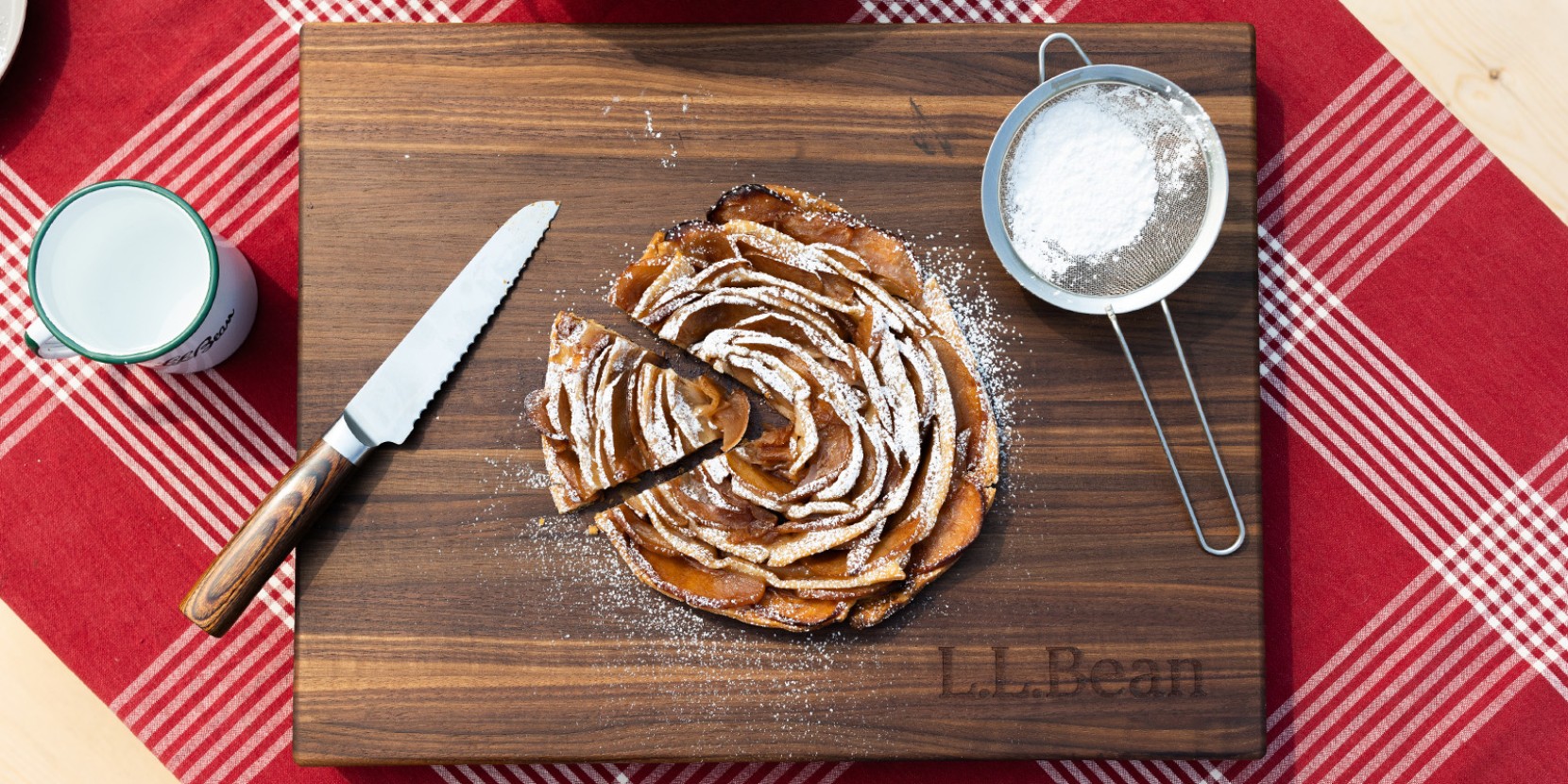A sliced apple pie on a wooden cutting board with a knife and powdered sugar sifter on a red checkered tablecloth.