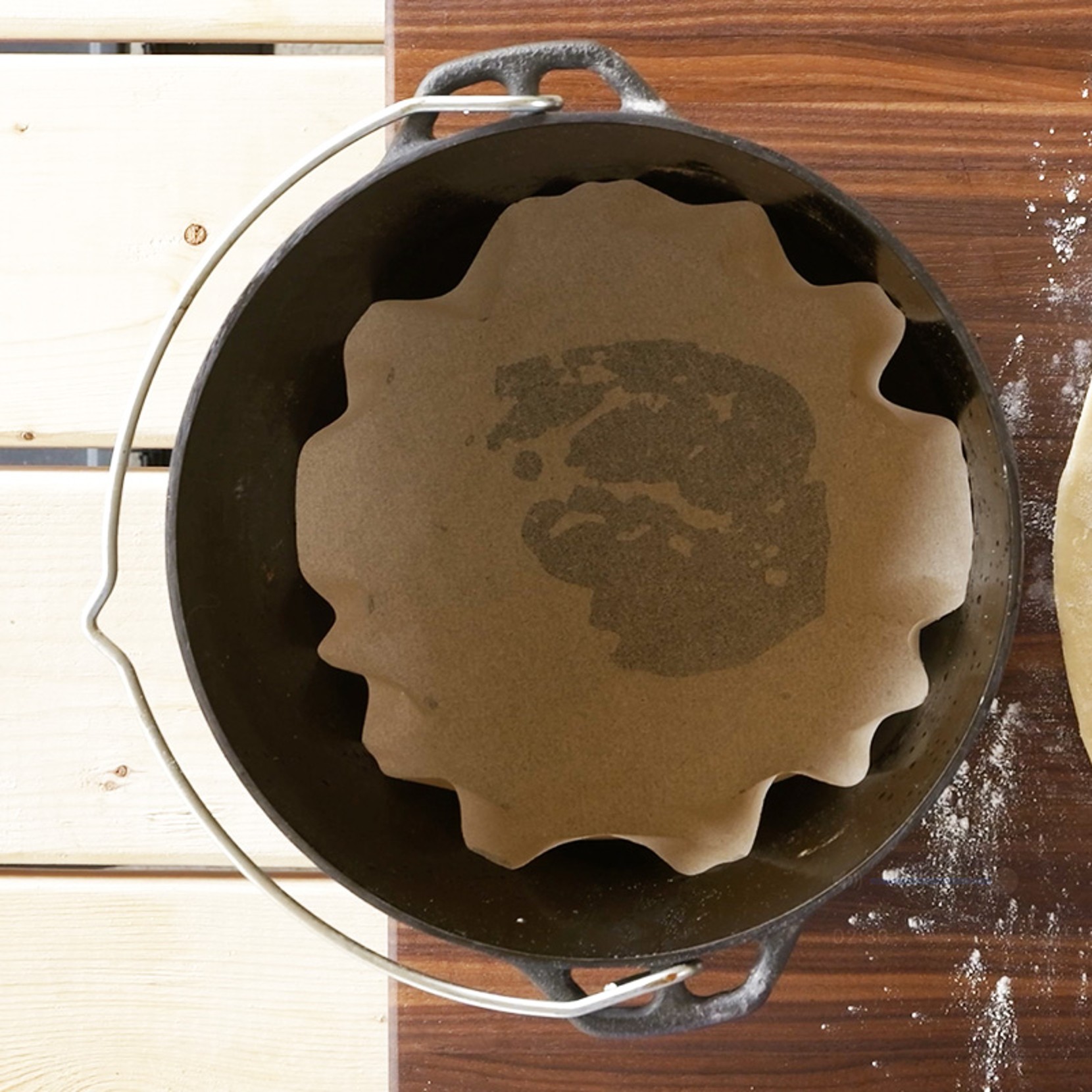 A round dutch oven lined with parchment paper on a wooden surface, with flour and dough nearby.