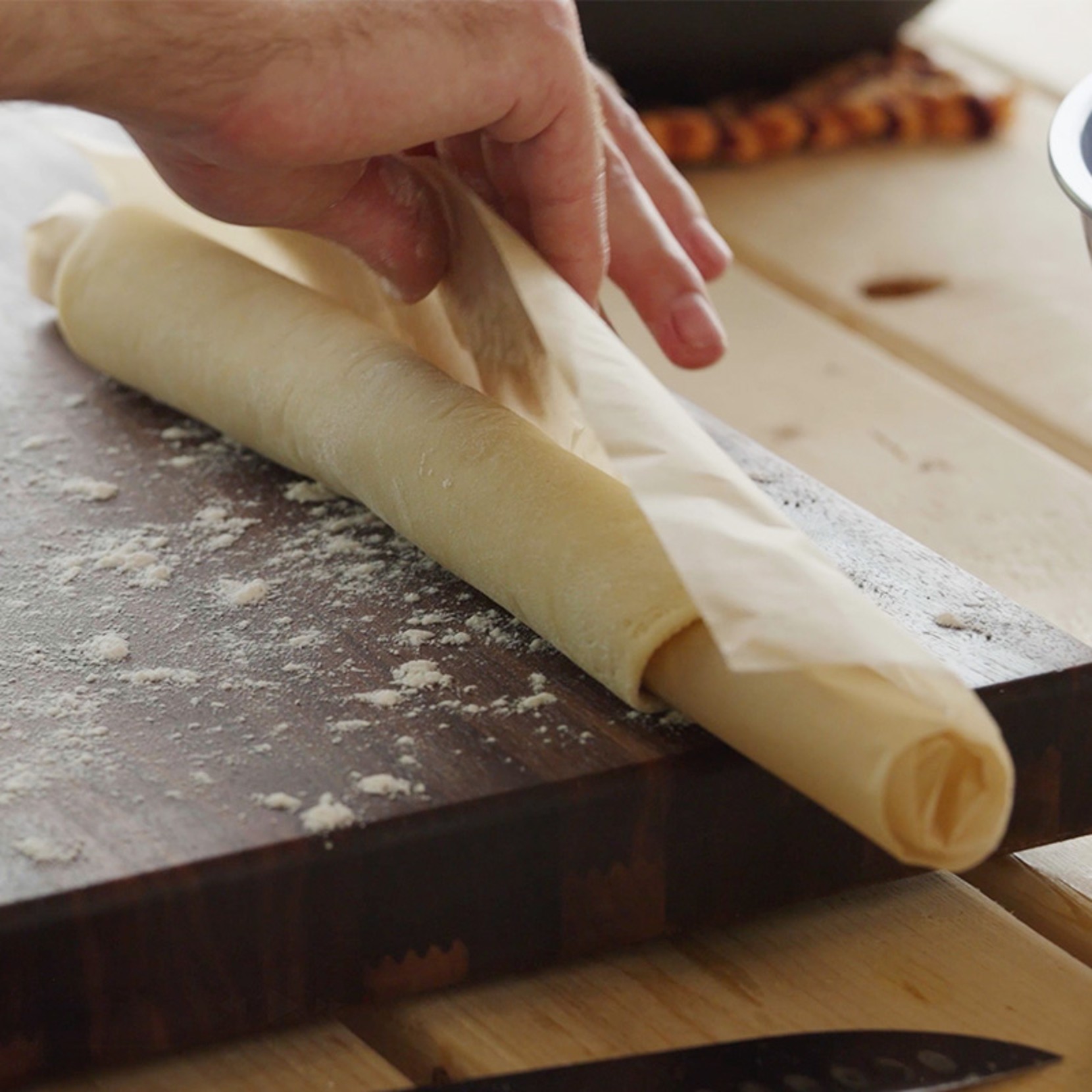 A hand rolls out a sheet of dough on a floured wooden cutting board.