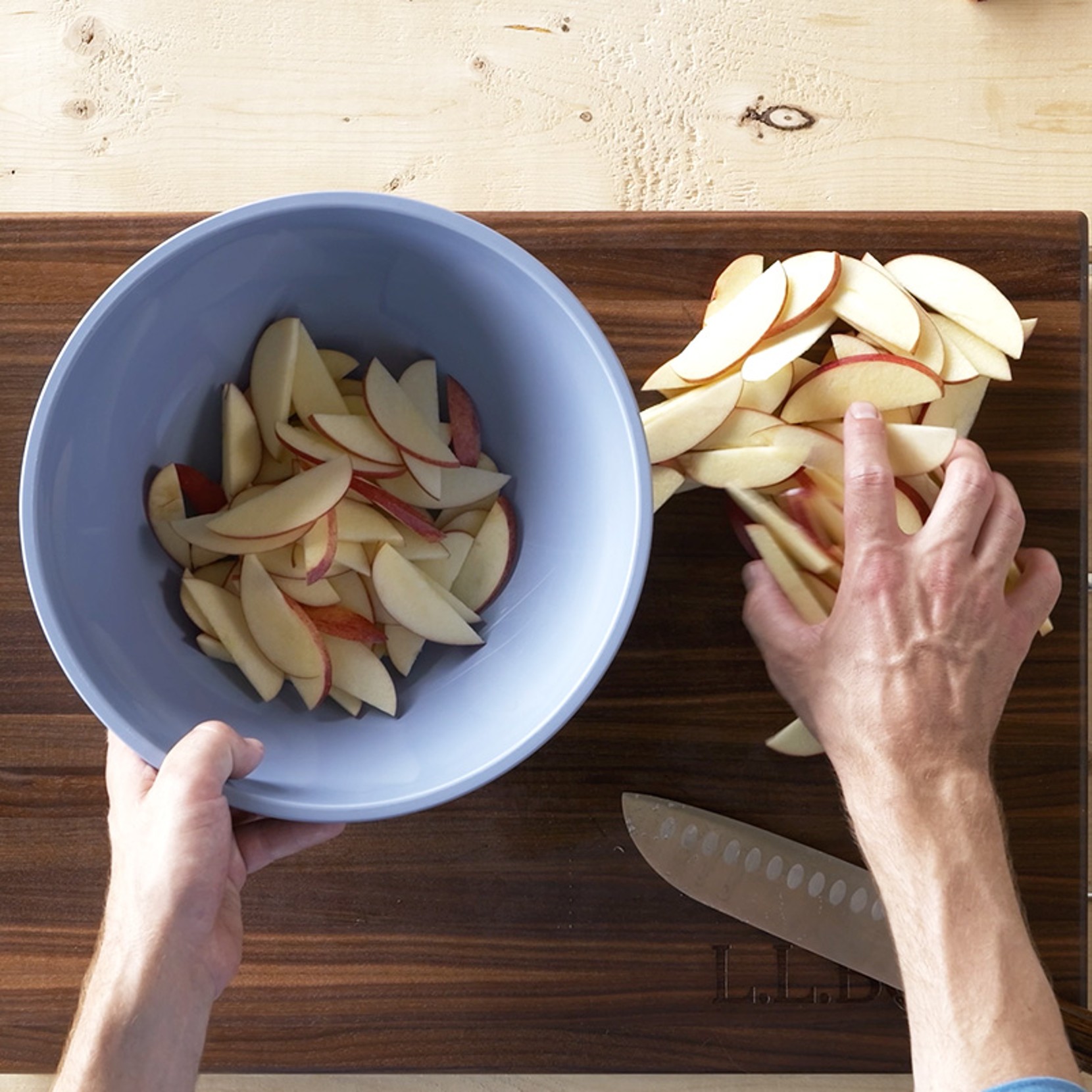 Two hands transfer thinly sliced red apples from a wooden cutting board into a blue bowl, with a knife nearby.