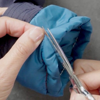 Hands use small scissors to cut a thread on the cuff of a blue and navy jacket sleeve.