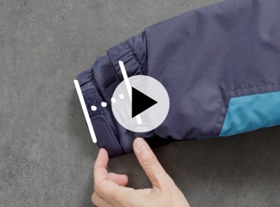 Hand adjusts Velcro strap on dark blue jacket cuff with teal accent; play button icon appears over image.