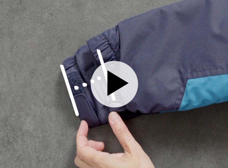 Hand adjusts Velcro strap on dark blue jacket cuff with teal accent; play button icon appears over image.