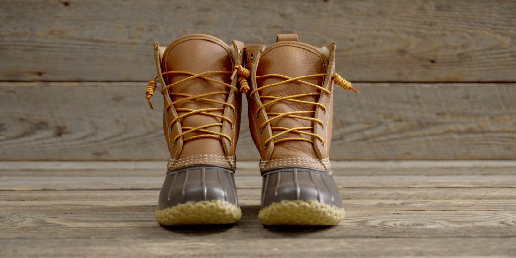 Brown leather duck boots with yellow laces and rubber soles, placed on a wooden surface.
