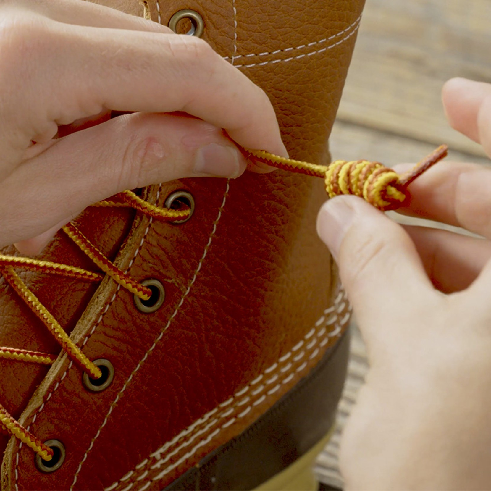 Hands tying yellow and red lace on a brown leather boot with metal eyelets and white stitching.