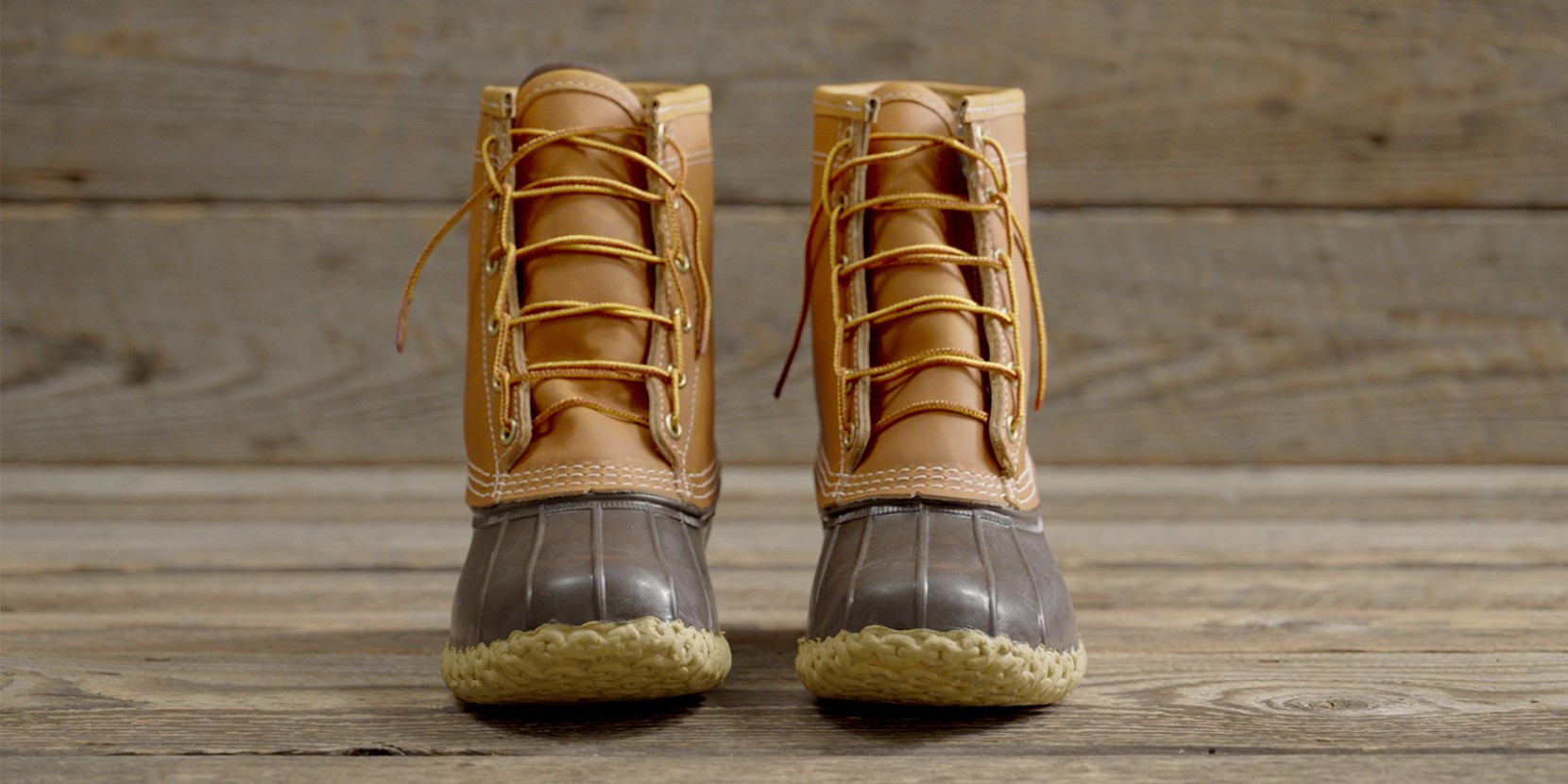 A pair of tan and brown duck boots with yellow laces on a wooden floor.