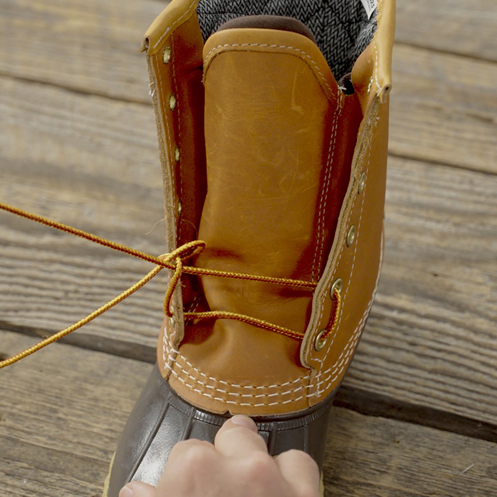 Hand tying laces on a brown leather boot with metal eyelets and white stitching, on a wooden surface.