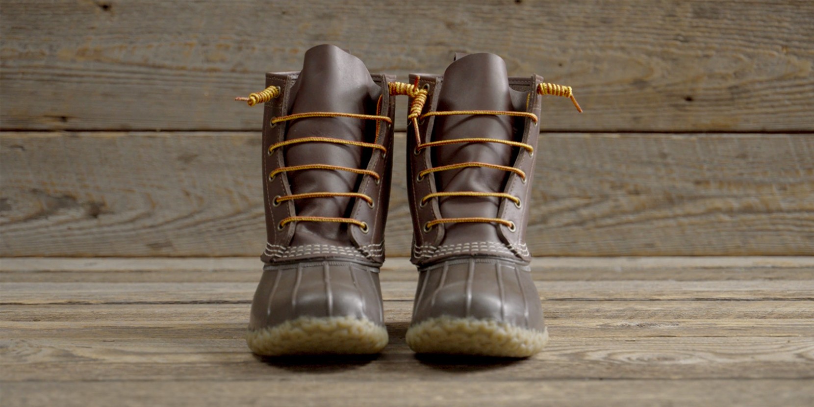 Pair of brown leather duck boots with yellow laces on a wooden surface and background.