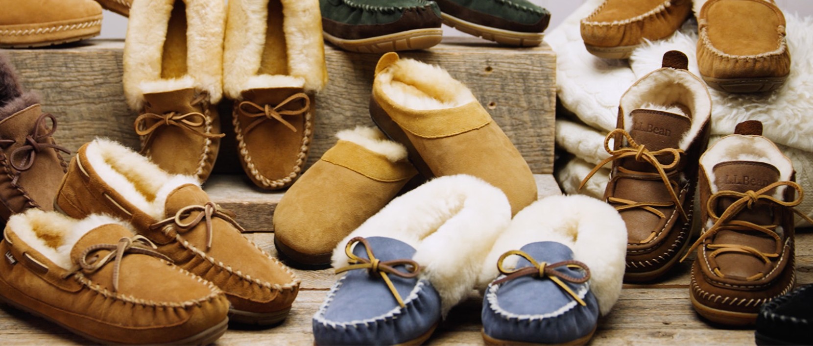 Assorted shearling-lined moccasin slippers in brown, tan, blue, and green displayed on rustic wooden shelves.
