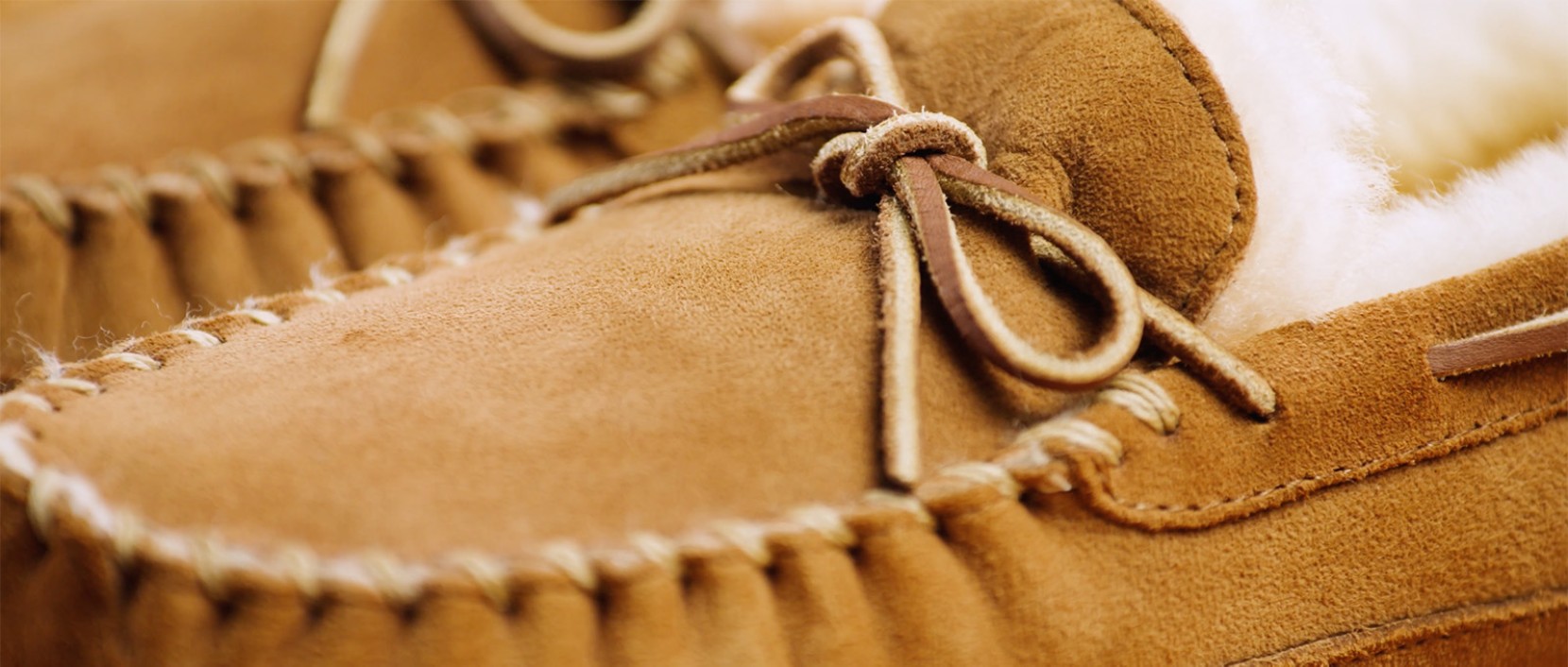 Close-up of a tan suede moccasin slipper with white fleece lining and a tied leather lace detail.